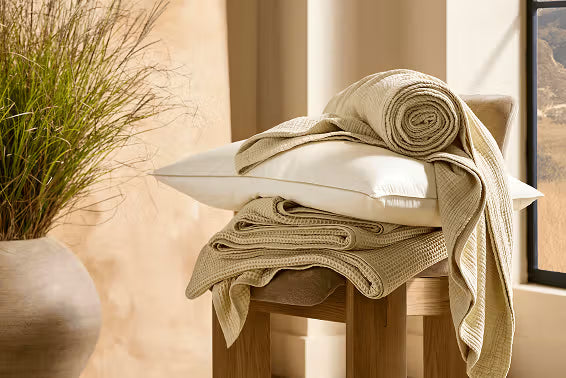 Folded beige towels on a wooden stool with a plant and window in the background